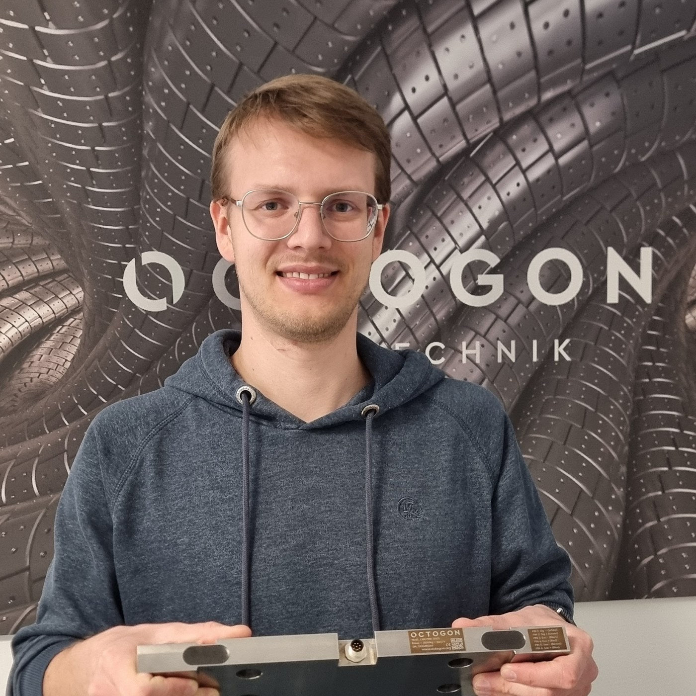 Mitarbeiter von octogon hält eine Biegestab-Wägezelle vor der octogon-Logowand